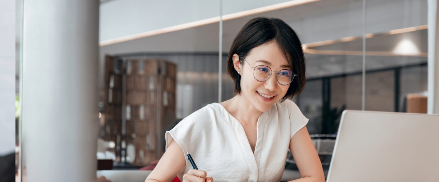 Young professional smiling while taking notes and using a laptop at a desk in a modern office workspace.